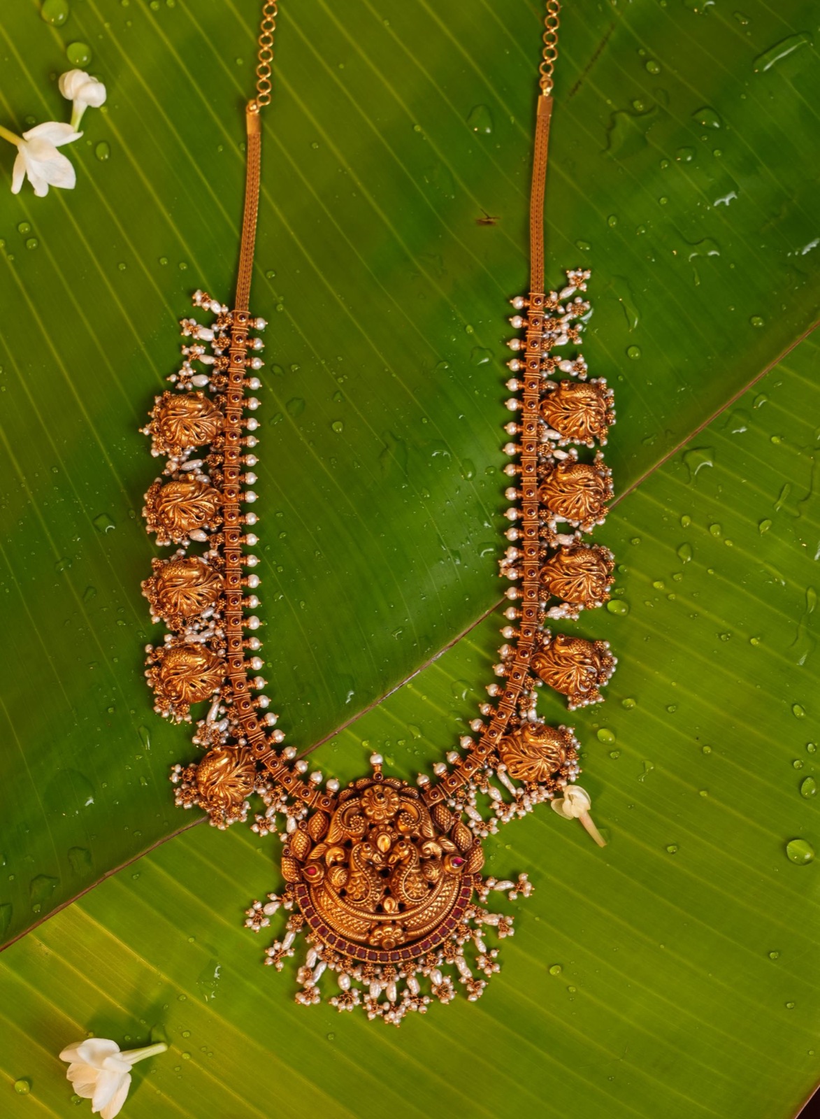 Antique gold temple necklace with peacock motifs, rubies and pearl clusters on a banana leaf with jasmine flowers